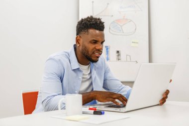 A focused young man sits at a desk, typing on his laptop with a mug and markers nearby. He is engaged in some work amidst charts on the whiteboard behind him. clipart