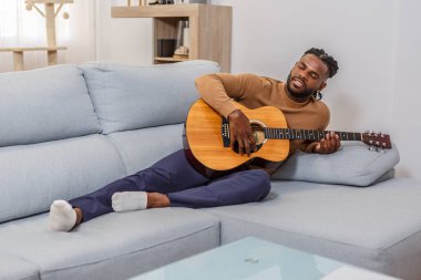 Bir adam açık renkli bir kanepede akustik gitar çalarak rahatça yaslanır..