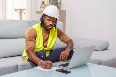 A construction worker in a yellow safety vest and hard hat sits on a couch, taking notes while looking at his laptop. The workspace is bright and inviting, suitable for productivity. clipart