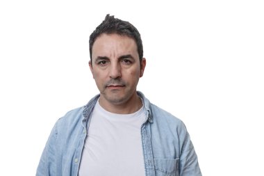 Beyaz bir gömleğin üstüne açık renk kot ceket giyen bir adam ciddi bir ifadeyle ayakta duruyor. Kısa siyah saçları var ve arka planı bembeyaz, özelliklerini vurguluyor..