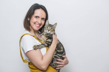 Kısa saçlı, parlak sarı bir tulum giymiş genç bir kadın çizgili kedisini sevgiyle tutarken gülümsüyor. Bu an, evcil hayvanlarla paylaşılan neşe ve sevgiyi yakalar..