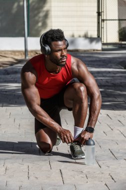 Canlı bir halk parkında, Afro-Amerikalı bir adam spor aktivitesine hazırlanırken bağcıklarını bağlamak için diz çöker. Spor aletleri ve kulaklık takıyor, fitness 'e olan bağlılığını gösteriyor..