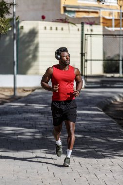 Afro-Amerikalı bir adam halka açık bir parkta kırmızı kolsuz gömlek ve siyah şort giyerek koşuyor. Kulaklıkla müzik dinler ve temiz havada çalışmaktan zevk alır..