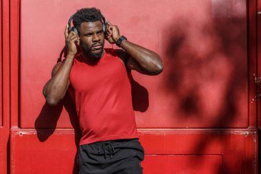 Yoğun çalışma seansını tamamladıktan sonra kulaklık takan bir adam canlı kırmızı bir duvara yaslanıyor. Rahatlamış ve müziğine dalmış görünüyor. Rahatlamak için biraz zaman ayırıyor..