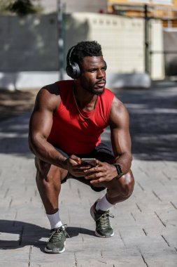 Kırmızı kolsuz tişörtlü ve atletik şortlu bir adam elinde akıllı bir telefonla parkta çömeliyor. Kulaklık takıyor ve odaklanmış görünüyor. Öğlen güneşinde antrenmanının keyfini çıkarıyor..