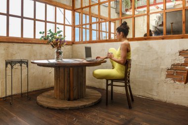 A yoga teacher sits in a sunny studio, sipping tea while organizing her class on a laptop. The inviting space features large windows and a simple wooden table. clipart