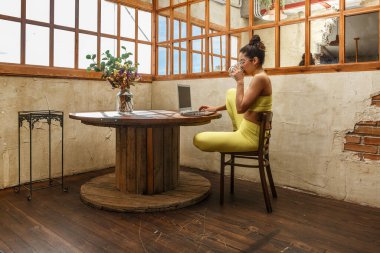 Yoga hocası parlak bir stüdyoda ahşap bir masada oturmuş çayını yudumluyor ve yaklaşan derse hazırlanırken bilgisayarına odaklanıyor..