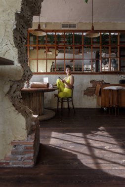 Canlı sarı giyinmiş yoga için hazırlanan bir kadın ahşap bir taburede oturuyor, elinde bir telefon tutuyor ve yüzünü yana çeviriyor. Güneş ışığı büyük pencerelerden süzülür, sıcak gölgeler yaratır..