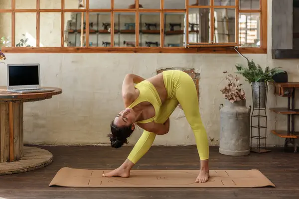 Kırsal ahşap döşemeli bir stüdyoda, sarı saçlı bir kadın yoga pozu verirken zarifçe dengeliyor. Yakınlarda bir dizüstü bilgisayar duruyor, dingin atmosferi arttırıyor..