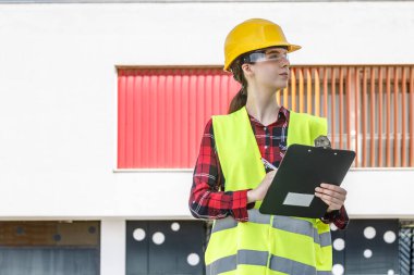 Sarı kasketli ve yansıtıcı yelekli bir kadın inşaat müfettişi binanın dışında duruyor ve proje ilerlemesini elinde bir panoyla değerlendiriyor..