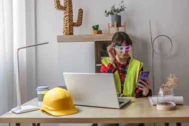 Çağdaş bir ofis ortamında, güvenlik yeleği giymiş bir kadın akıllı telefonunu kontrol ederken yapay zeka gözlüğünü akıllıca ayarlar..