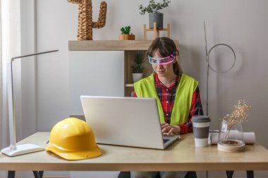 Şık bir ofis ortamında bir kadın dizüstü bilgisayarında yazarken yapay zeka gözlüğü kullanıyor. Güvenlik yeleği giymiş, teknolojinin ve modern çalışma alanı estetiğinin bir karışımını sergiliyor..