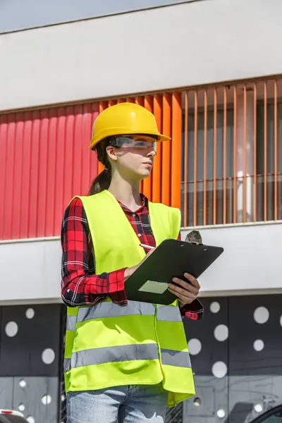 Sarı bir şapka ve güvenlik yeleği giyen kadın bir inşaat işçisi, elinde bir not panosuyla çağdaş bir binanın önünde güvenle duruyor. Araziyi inceliyor, güvenliğin sağlanması için..