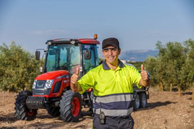 Sarı ve gri üniformalı, gülümseyen bir çiftçi, bir zeytin koruluğunda gururla durur, kırmızı bir traktörün yanında başparmağını kaldırır..