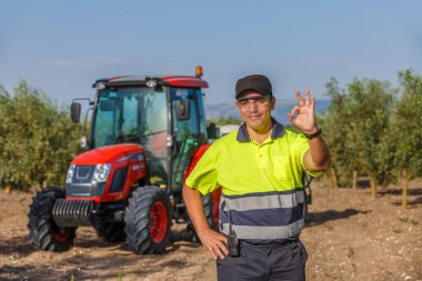 Parlak güvenli kıyafetler giymiş neşeli bir çiftçi zeytin korusunda duruyor ve iyi bir jest işareti veriyor. Yakınlarda bir kırmızı traktör var, açık mavi gökyüzünün altındaki tarımsal ortamı vurguluyor..