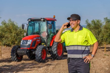 Güvenlik üniformalı bir çiftçi zeytin koruluğunda duruyor, cep telefonuyla yakınlardaki kırmızı bir traktörle konuşuyor..