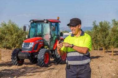 Parlak sarı gömlekli bir traktör operatörü verimli bir tarım alanında akıllı telefonunu kontrol ederken gülümsüyor. Arka planda, çiftçilik ortamını vurgulayan kırmızı bir traktör görünür..