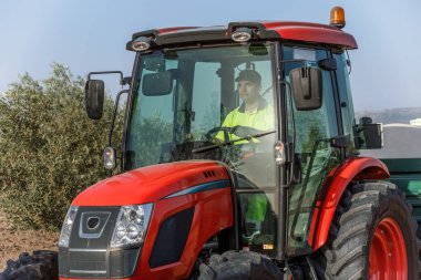 Bir çiftçi, açık mavi gökyüzünün altında ve yeşilliklerle çevrili tarlalardan geçen parlak kırmızı bir traktör kullanır. Sahne, modern tarım ve kırsal yaşamın özünü yakalıyor..