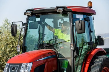 Parlak sarı tişörtlü bir çiftçi kırmızı bir traktör kullanıyor, zeytin koruluğundan geçiyor. Berrak gökyüzü ve yemyeşil ağaçlar, günlük tarım faaliyetlerini gösteren sakin bir tarım atmosferi yaratıyor..