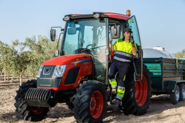 Bir erkek çiftçi, zeytinlikte kırmızı bir traktörün yanında duruyor, tarımsal görevlerine başlamaya hazır..