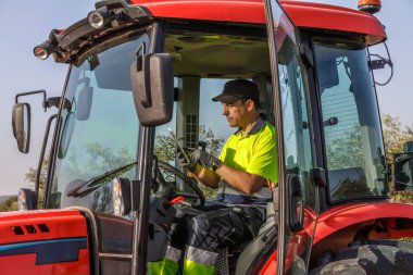 Endülüs 'te güneşli bir zeytinlikte bir traktörde uzman bir çiftçi tablet kullanıyor. Arazide dikkatli bir şekilde geziniyor. Hasatın başarısını uzman ellerle ve kararlı bir şekilde sağlıyor..