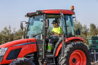 Profesyonel bir çiftçi zeytinlikte traktör kullanıyor, tabletteki verileri kontrol ediyor. Endülüs 'ün manzaralı arkaplanı modern tarım uygulamalarını tamamlıyor.
