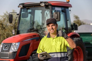 Güler yüzlü bir çiftçi, elinde bir pano, elinde kırmızı bir tarımsal alanda kırmızı bir traktörün yanında duruyor..