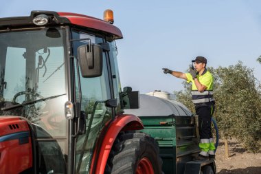 Yüksek görüşlü gömlekli bir işçi, tarımsal bir alanda traktör kullanırken telsizle iletişim kurar..