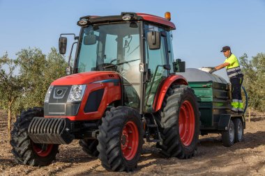 Bir çiftçi, gübre tankıyla donatılmış kırmızı bir traktörü kullanıyor. Titizlikle parlak güneş ışığı altında bir zeytin koruluğuna mahsul püskürtüyor..