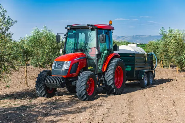 İspanya 'nın Endülüs kentindeki bir zeytinlikte çalışan parlak kırmızı bir traktörün römork çektiği görülüyor. Manzara, açık mavi gökyüzünün altında yemyeşil zeytin ağaçlarını barındırıyor..
