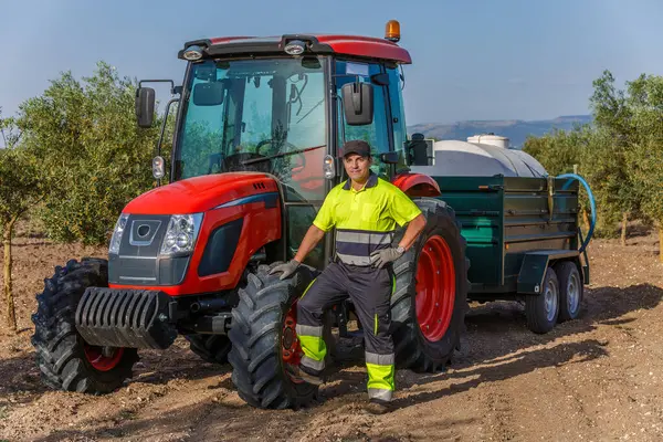 Bir tarım işçisi, açık mavi gökyüzünün altında zeytin ağaçlarıyla çevrili bir su tankı ile donatılmış modern bir kırmızı traktörün yanında poz veriyor. Kurulum, tarımsal yaşamın ve ekipmanların özünü yakalıyor..