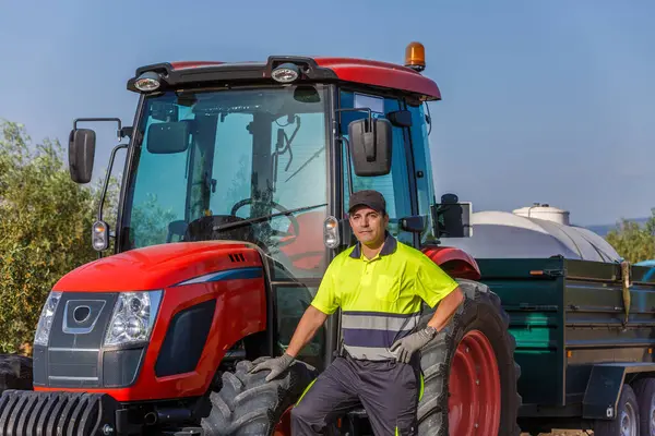 Yetenekli bir traktör sürücüsü Endülüs 'te güneş ışığında bir zeytinlikte kırmızı traktörünün yanında duruyor. Güvenlik yeleği giyiyor ve tarıma olan bağlılığını göstermek için toprağı işlemeye hazır..
