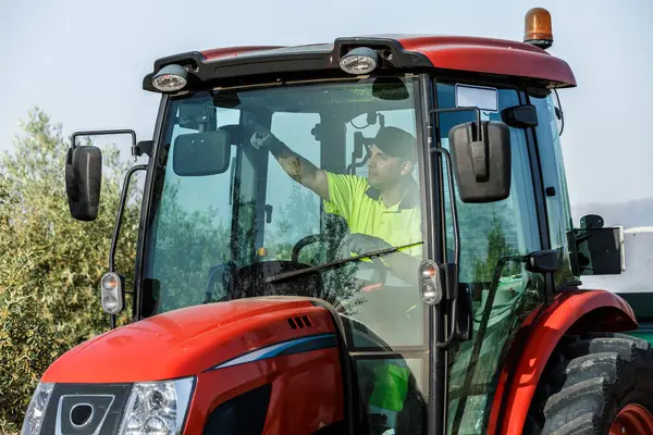 Parlak sarı tişörtlü bir çiftçi kırmızı bir traktör kullanıyor, zeytin koruluğundan geçiyor. Berrak gökyüzü ve yemyeşil ağaçlar, günlük tarım faaliyetlerini gösteren sakin bir tarım atmosferi yaratıyor..