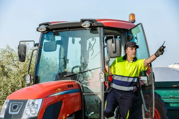 Bir çiftçi kırmızı traktöründen Endülüs 'te güneşli bir zeytin koruluğunda çıkar ve bir telsizle el kol hareketi yapar. Zeytin toplama, geleneksel tarım uygulamalarını sergileme sürecinde yer alıyor..