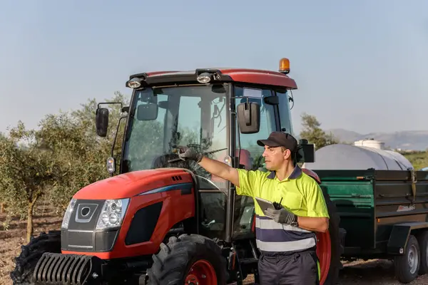 Neon gömlek ve eldiven giyen neşeli bir çiftçi yemyeşil zeytin ağaçlarının arasında kırmızı bir traktör kullanıyor. Tarımsal çalışmanın ve çiftçilikteki neşenin özünü yansıtıyor..