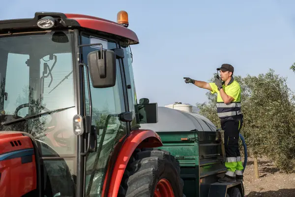 Yüksek görüşlü gömlekli bir işçi, tarımsal bir alanda traktör kullanırken telsizle iletişim kurar..