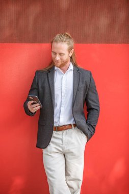 Şık, kızıl saçlı, uzun saçlı işadamı akıllı telefonunu kullanırken kırmızı duvara dayanıyor. Spor ceket giyiyor ve rahat ve profesyonel görünüyor..