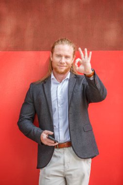 Renkli bir duvarın önünde duran kızıl saçlı işadamı akıllı telefonunu ve el hareketlerini tutuyor. Ceket giyiyor ve mutlu görünüyor..