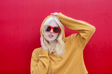 Genç bir albino kadın, kırmızı gözlüklü, sarı bir kapüşonlu, burnuna tünemiş, canlı kırmızı bir arka plan üzerine kurulmuş duruyor..