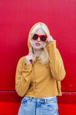 Albinizme sahip genç bir kadın cesur bir kırmızı zemine karşı özgüvenle poz veriyor. Kapüşonlu bir kazak ve şık bir güneş gözlüğü takıyor. Modern ve modaya uygun bir görünüşü var..