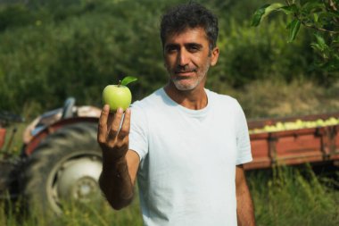 Elinde elmayla arka planda traktörle elma bahçesinde poz veren bir ortadoğunun portresi..