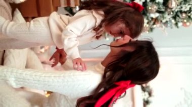 Anne ve kızı, beyaz elbiseli ve saçlarında kırmızı kurdeleyle bir Noel ağacının önünde dinleniyorlar..