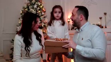 Şaşırtıcı duyguları olan mutlu bir aile hediye kutusunu açarken arka planda bir Noel ağacı, şömine ve mumlar var..