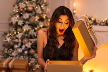 Bir hediye kutusunu açarken sürpriz duyguları olan esmer bir kadının portresi. Arka planda bir Noel ağacı ve şömine var..