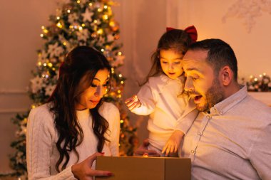 Şaşırtıcı duyguları olan mutlu bir aile hediye kutusunu açarken arka planda bir Noel ağacı, şömine ve mumlar var..