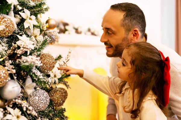 Portrait of a father and daughter looking at a christmas tree with a smile on their faces in front of a fireplace.