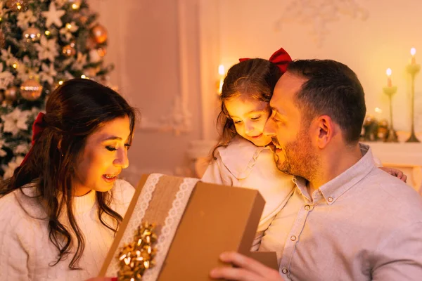 Şaşırtıcı duyguları olan mutlu bir aile hediye kutusunu açarken arka planda bir Noel ağacı, şömine ve mumlar var..