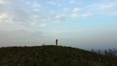 Drone footage of a 30s one woman with red tights and tops dancing on a Tumulus hill on a cloudy sky background