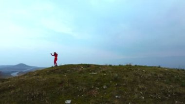 Drone footage of a 30s one woman with red tights and tops dancing on a Tumulus hill on a cloudy sky background