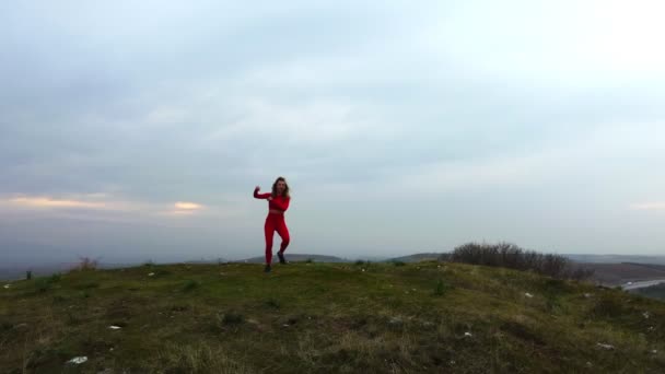 30代のドローン映像では 曇り空の背景に赤いタイツとトップスを着た女性が古墳の丘で踊っている — ストック動画